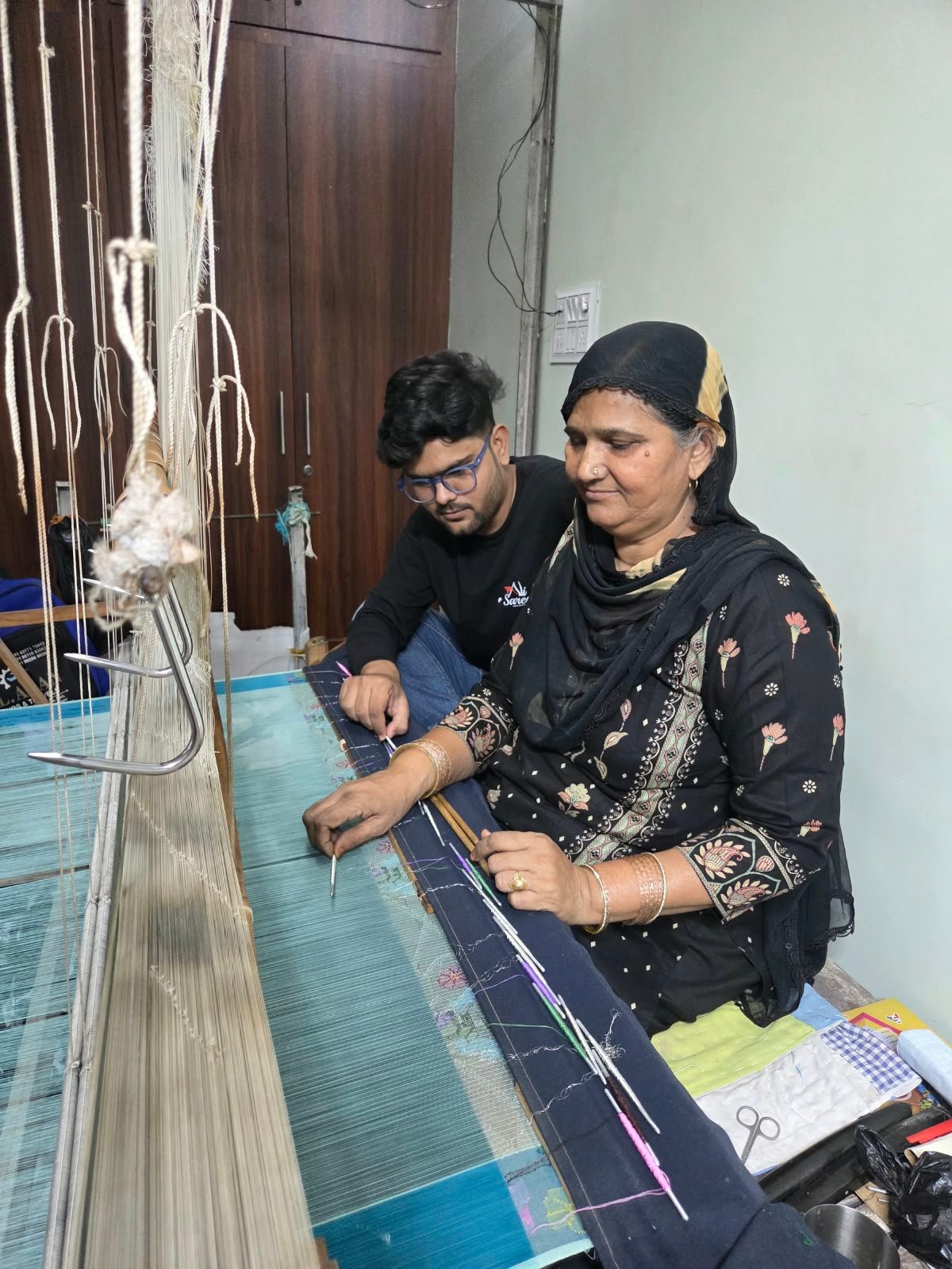 A skilled female artisan wearing a black floral kameez and headscarf works at a traditional handloom, intricately weaving fine textile threads alongside a young man in glasses observing and assisting the process.